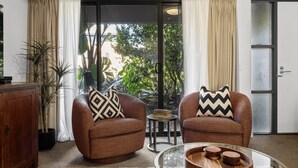 Una habitación con dos sillones marrones, una mesita redonda, un aparador y un ventanal corredero que da a un jardín.