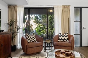 A room with two brown armchairs, a small round table, a cabinet, and a sliding glass door leading to a garden.
