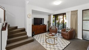 Una sala de estar con una escalera, un televisor de pantalla plana, dos sillones, una mesa de centro y un ventanal con cortinas.