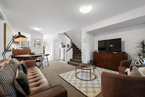 A modern living room with a brown leather sofa, a dining table with chairs, a staircase, a television on a wooden cabinet, and a patterned rug.