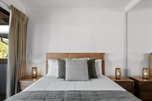 Un lit avec une tĂȘte de lit en bois, deux tables de chevet avec des lampes et une vue sur la verdure Ă lâextĂ©rieur.