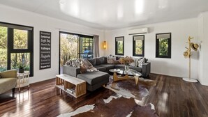 Een ruime woonkamer met een grote grijze hoekbank, een glazen salontafel en een houten bijzettafeltje. Er zijn grote ramen die natuurlijk licht en uitzicht op de omgeving bieden.