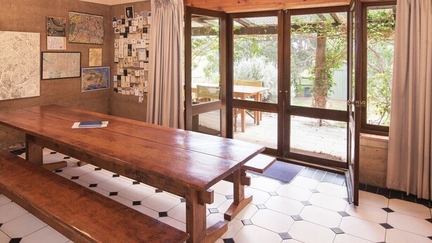A room with a wooden table and benches, a wall with various framed items, and a large glass door leading to an outdoor area with a table and chairs.