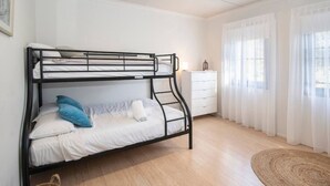 Una cama litera con ropa de cama blanca, un aparador blanco y una alfombra tejida sobre piso de madera.