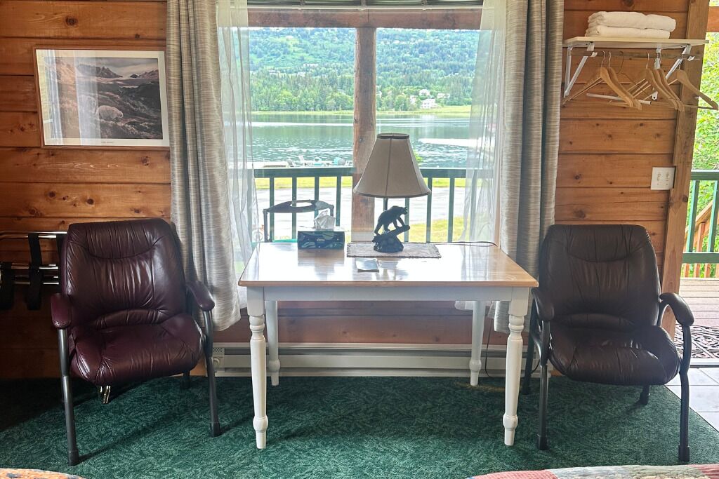 A room with a wooden interior, a table with a lamp, two leather chairs, a view of a lake, and a hanging rack with towels.