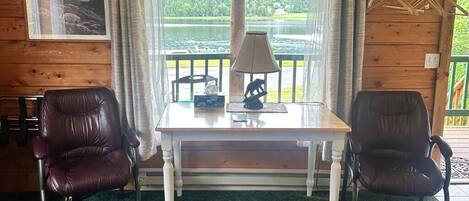 A room with a wooden interior, a table with a lamp, two leather chairs, a view of a lake, and a hanging rack with towels.