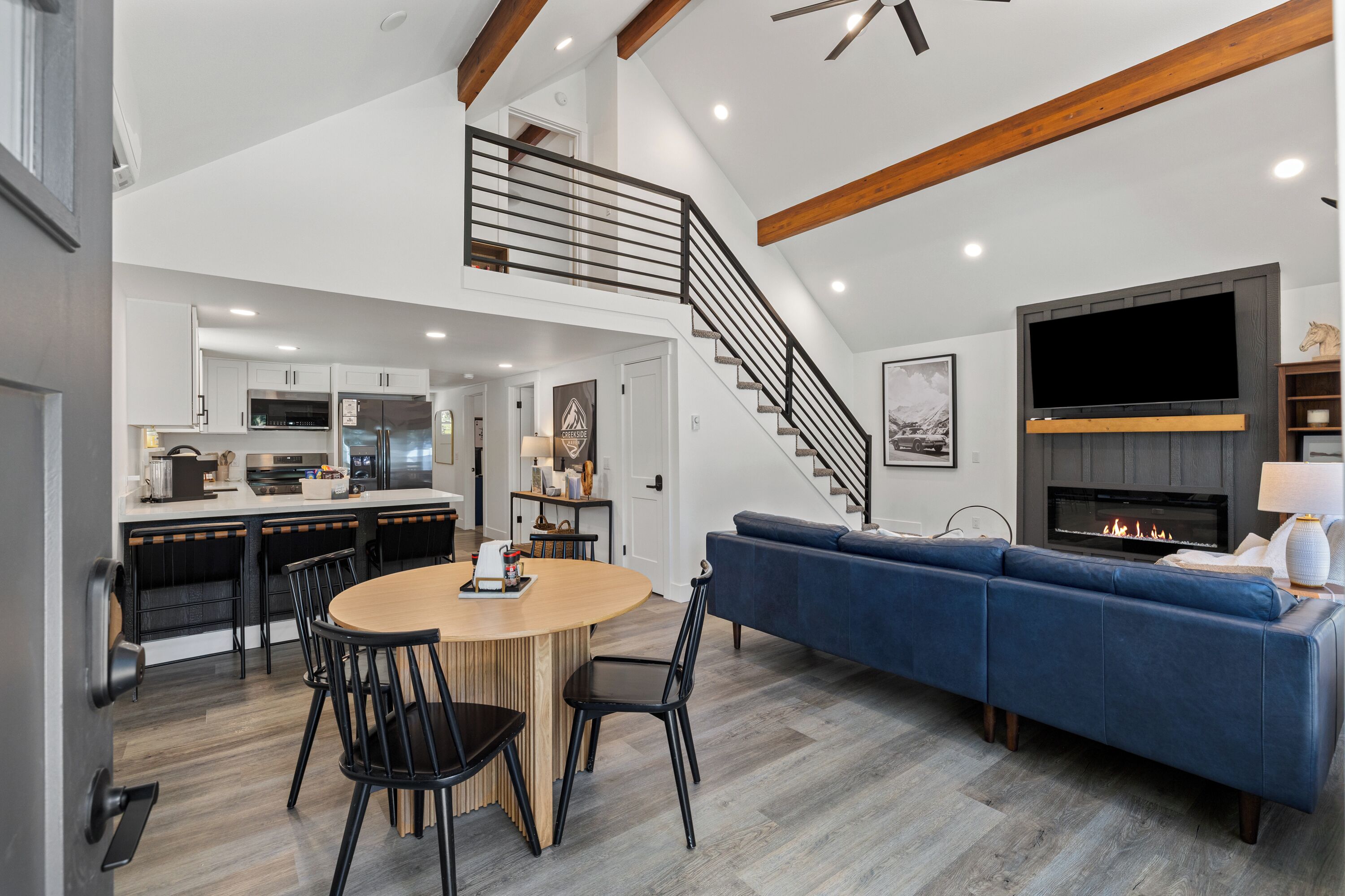 A modern living room with a blue sofa, a round wooden dining table with chairs, a kitchen with white cabinets, and a staircase leading to an upper level.