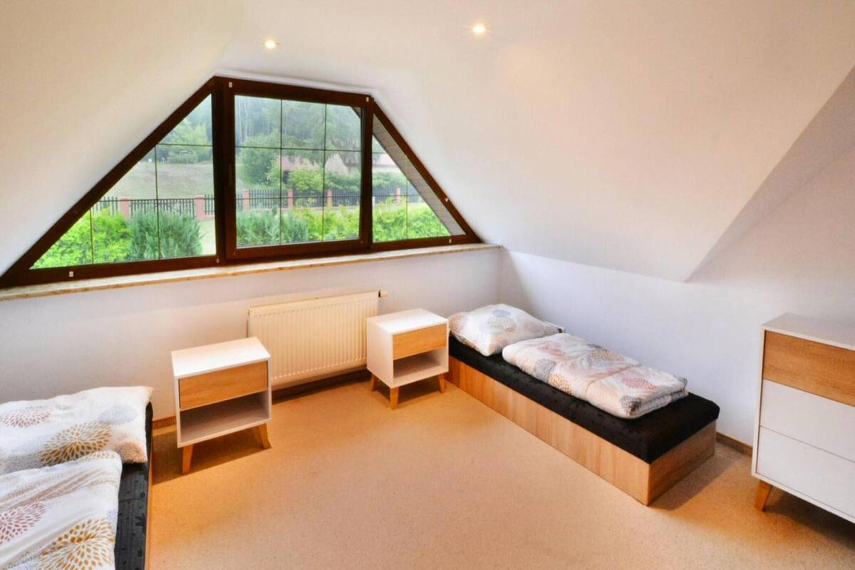 A bedroom with a sloped ceiling, a bed, a bedside table, a small desk, and a window with a view of greenery.