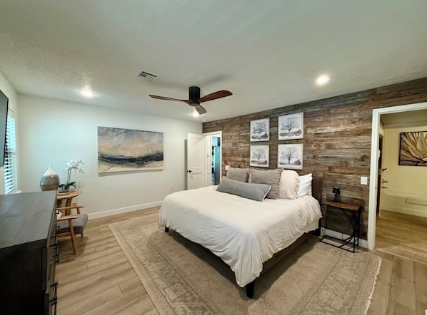 A bedroom with a bed, a ceiling fan, a wooden accent wall, and a door leading to another room.
