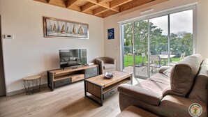 A modern living room with a TV, sofa, and coffee table. There is a painting of sailboats on the wall and a view of the garden through large windows.