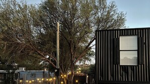 Un área de grava con una cerca metálica, un pequeño edificio con una ventana y árboles al fondo.