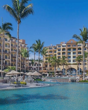 Un hotel con varios pisos, un área de piscina con sillas y sombrillas, y palmeras.