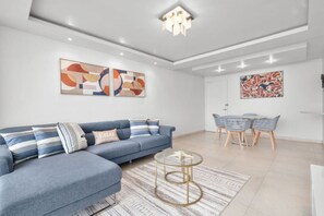 Un salon moderne avec un canapé d’angle bleu, une table basse en verre et un coin repas avec des chaises en osier.