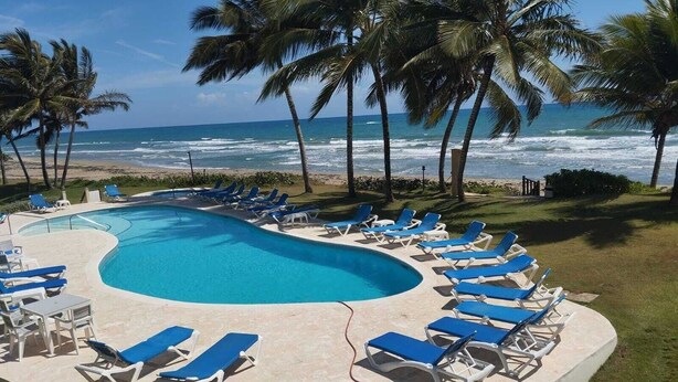A pool area with blue lounge chairs, a curved pool, and a view of the ocean with palm trees. - A-2 Oceanfront at The Victorian (Cabarete)