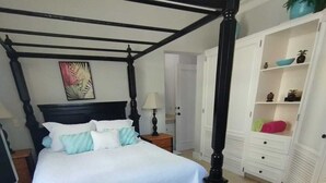 A four-poster bed with a white bedspread and teal pillows, a nightstand with a lamp, and a white wardrobe with open doors revealing shelves and drawers. - A-2 Oceanfront at The Victorian (Cabarete)