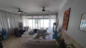 A spacious living room with a large sofa, a dining area with a table and chairs, ceiling fans, and large windows with curtains. - A-2 Oceanfront at The Victorian (Cabarete)