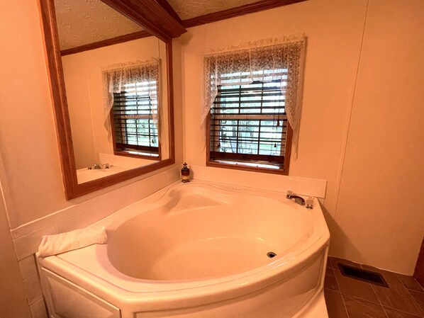 A bathroom with a large bathtub, a mirror, and two windows with lace curtains.