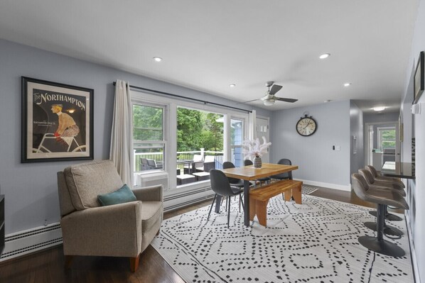 A modern living room with a sofa, dining table, and chairs. There is a large window with a view of greenery outside.
