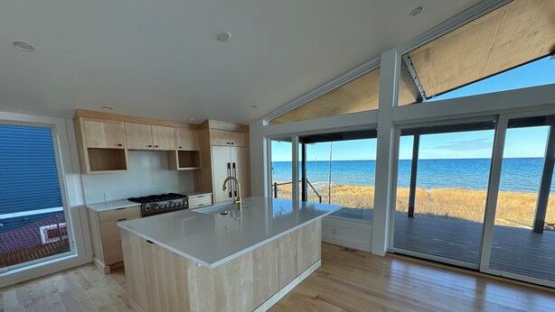 A modern kitchen with wooden cabinets, a central island, and a view of the ocean through large sliding glass doors. - Modern Lake Michigan Private Beach Cottage (Harbor Springs)