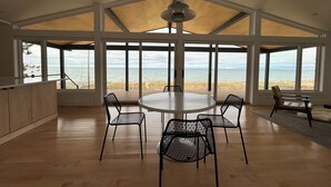 A spacious dining area with a large table and chairs, large windows offering a view of the beach and ocean, and a high ceiling with a skylight. - Modern Lake Michigan Private Beach Cottage (Harbor Springs)