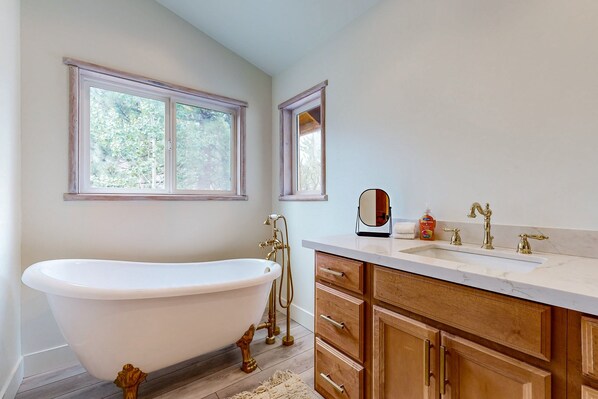 A bathroom with a freestanding bathtub, wooden cabinets, a marble countertop, and two windows with views of greenery.