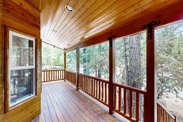 A wooden deck with a railing, large windows, and a view of trees.