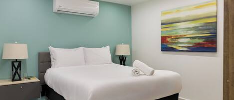 A modern bedroom with a bed, two bedside tables with lamps, an air conditioning unit, and a colorful abstract painting on the wall.