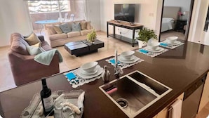 Une cuisine moderne dotée d’un îlot central, un coin repas avec un canapé et une table basse, et un salon avec un téléviseur et des fauteuils.
