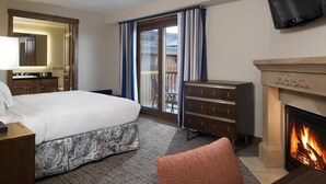 A hotel room with a bed, dresser, television, fireplace, and a balcony with curtains.