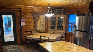 Ein Holzinterieur mit Kücheninsel, Esstisch, Stühlen, einem Kühlschrank und einem Fenster mit Vorhängen.