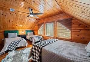 Une chambre mansardée chaleureuse avec des murs en bois, un plafond en pente, deux lits avec une literie à carreaux, une tête de lit recouverte d’un tissu vert et un ventilateur de plafond.