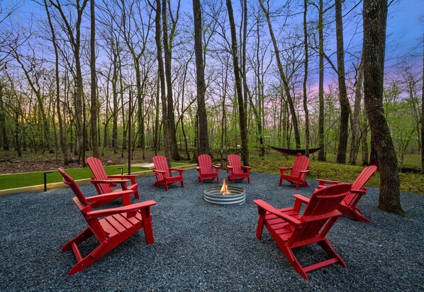 Un aménagement circulaire avec des chaises Adirondack rouges disposées autour d’un foyer central, le tout situé dans une zone boisée.