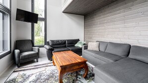 A modern living room with a grey sofa, a wooden coffee table, and a grey armchair. A flat-screen TV is mounted on the wall, and there is a window providing natural light.