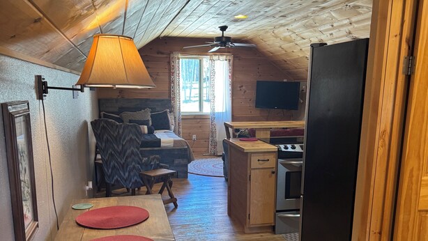 A cozy cabin interior with a wooden dining table, a bench, a ceiling fan, a kitchen area with a refrigerator, and a window with curtains.