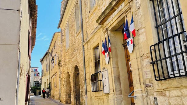 Une rue étroite bordée de bâtiments en pierre, de drapeaux et d’une enseigne indiquant « HÔTEL DE VILLE ».