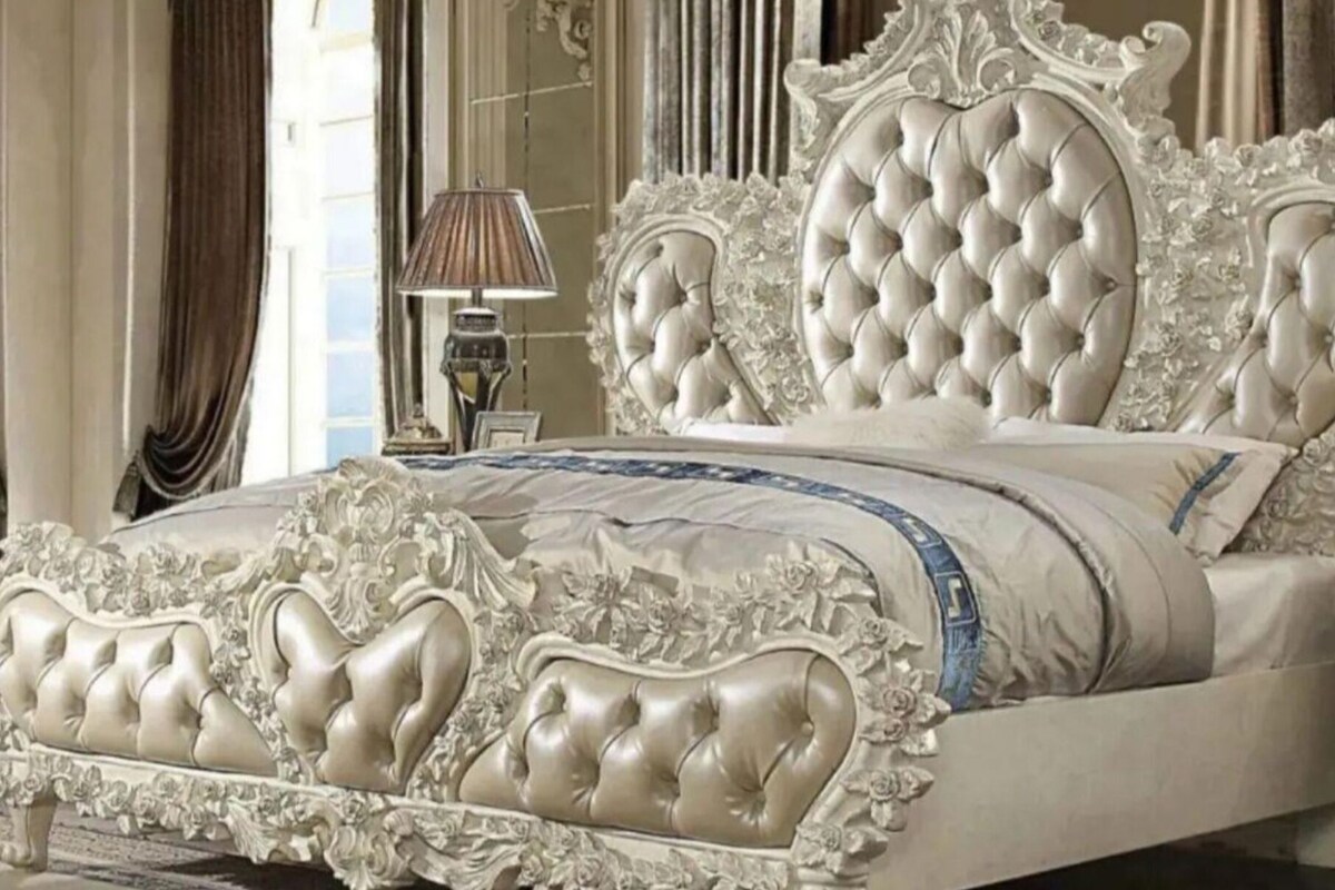 A luxurious bed with an ornate headboard and footboard, a bedside table with a lamp, and a view of a window with curtains.