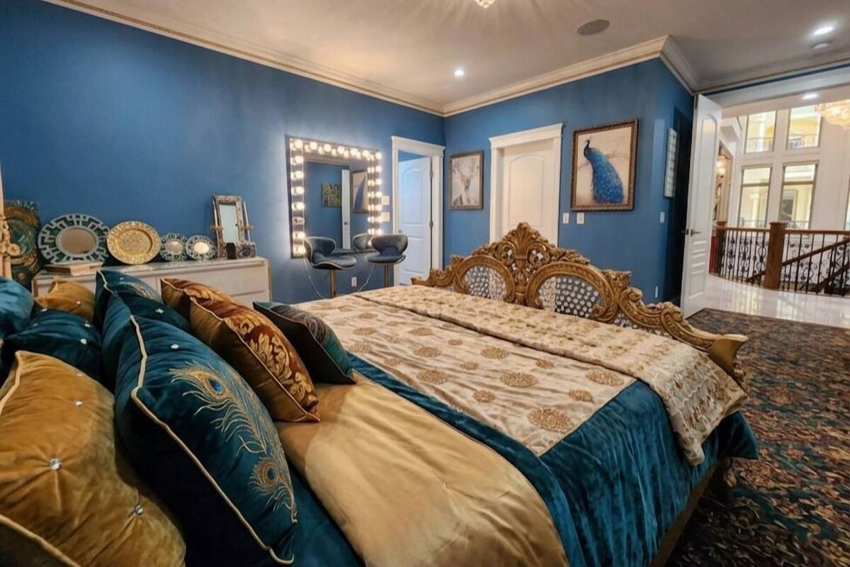 Een slaapkamer met een groot bed, een kaptafel met verlichting, een schilderij van een pauw en een trap naar een andere kamer.