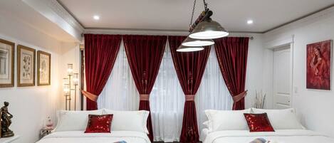 Habitación de hotel con dos camas, cortinas rojas, una araña y cuadros en las paredes.
