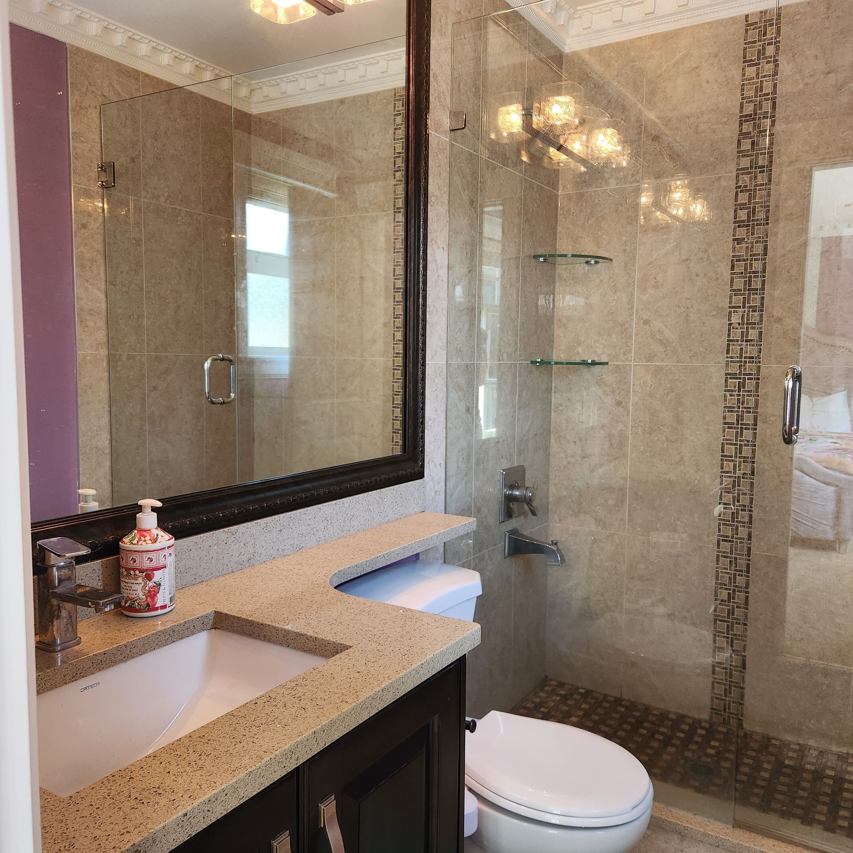 A bathroom with a glass-enclosed shower, a large mirror, a sink with a soap dispenser, and a toilet.