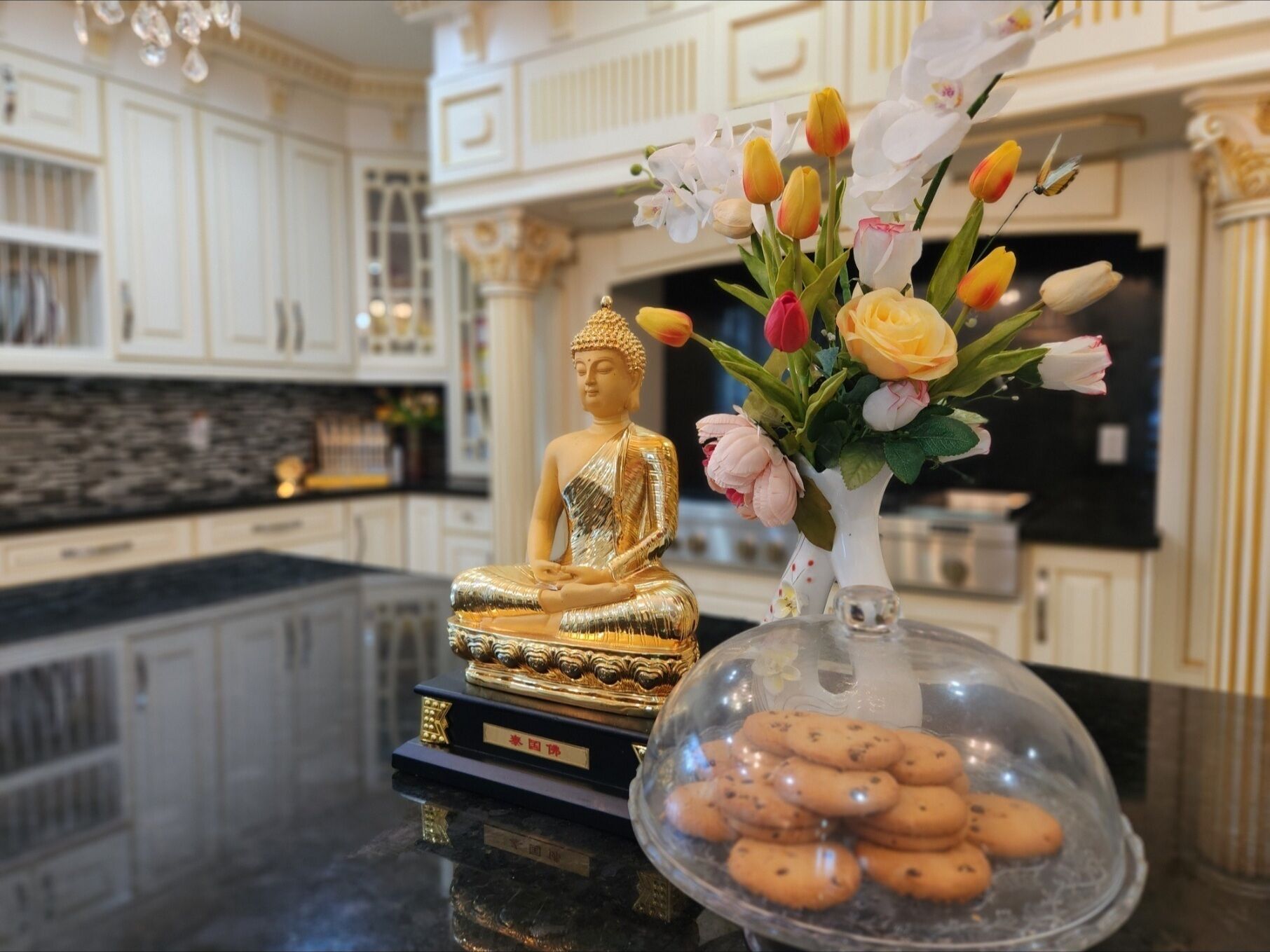 Une statue de Bouddha sur un piédestal, un vase rempli de fleurs et un plat de biscuits couvert, le tout posé sur un comptoir de cuisine.