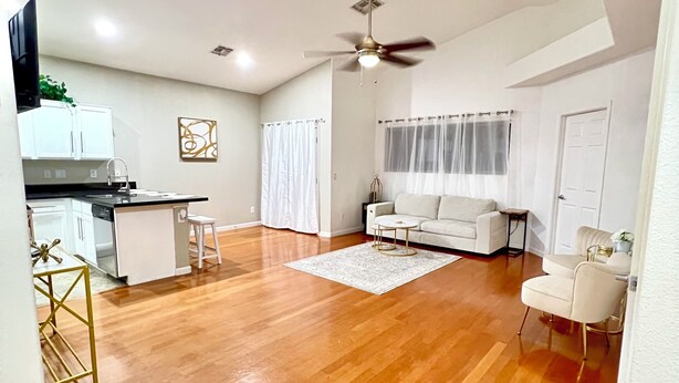 A spacious living area with a kitchen, white walls, wooden flooring, a ceiling fan, and a white sofa.