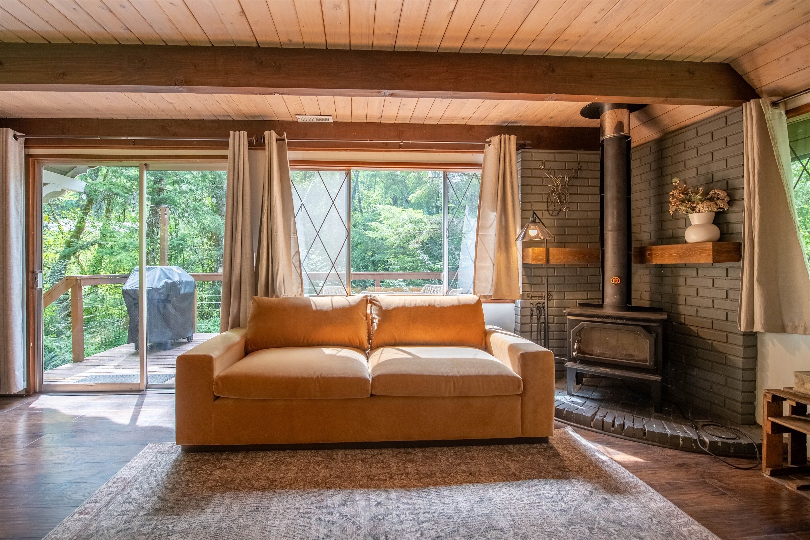 Een gezellige kamer met een houten plafond, een bank, een open haard en een groot raam met uitzicht op groen.