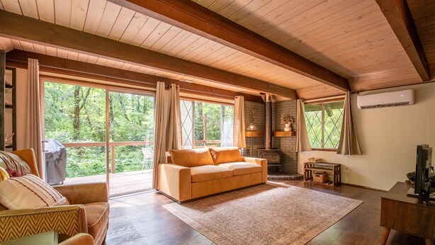 A cozy living room with a wooden ceiling, large windows, a sofa, a fireplace, and a TV stand. - Fog & Fern Hideaway - Cozy Cabin w/ Private Hot Tub near Mt. Rainier (Ashford)