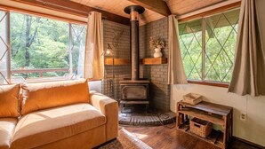 A cozy cabin interior with a wooden ceiling, a stone fireplace, a cushioned sofa, and large windows offering a forest view. - Fog & Fern Hideaway - Cozy Cabin w/ Private Hot Tub near Mt. Rainier (Ashford)
