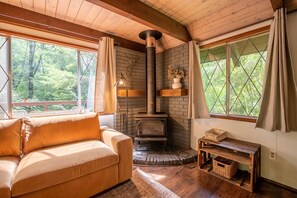 A cozy cabin interior with a wooden ceiling, a stone fireplace, a cushioned sofa, and large windows offering a forest view. - Fog & Fern Hideaway at Mt. Rainier (Ashford)