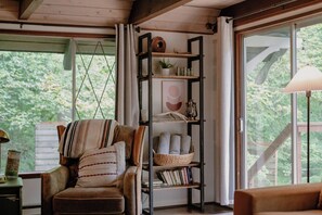 A cozy room with a leather armchair, a wooden bookshelf, and a large window with a view of greenery. - Fog & Fern Hideaway - Cozy Cabin w/ Private Hot Tub near Mt. Rainier (Ashford)