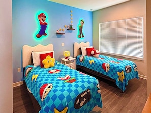 Et soveværelse med to senge, hver med et dynedækken med Mario-tema, en væg med billeder af Mario og Luigi, samt et vindue med persienner.