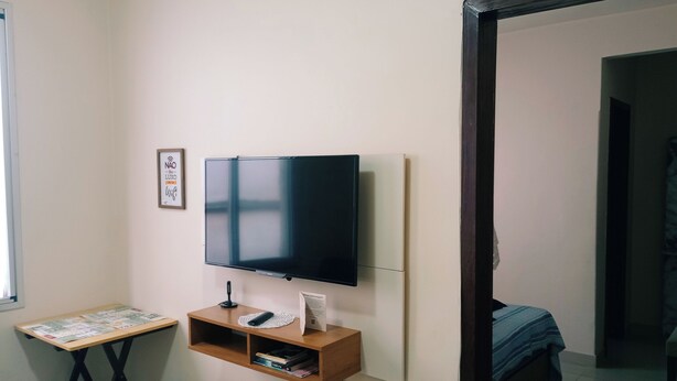 A room with a TV mounted on the wall, a small wooden desk with a chair, and a bed with a blue blanket. - Valença Space - in the heart of the historic center (Centro Histórico Porto Alegre)