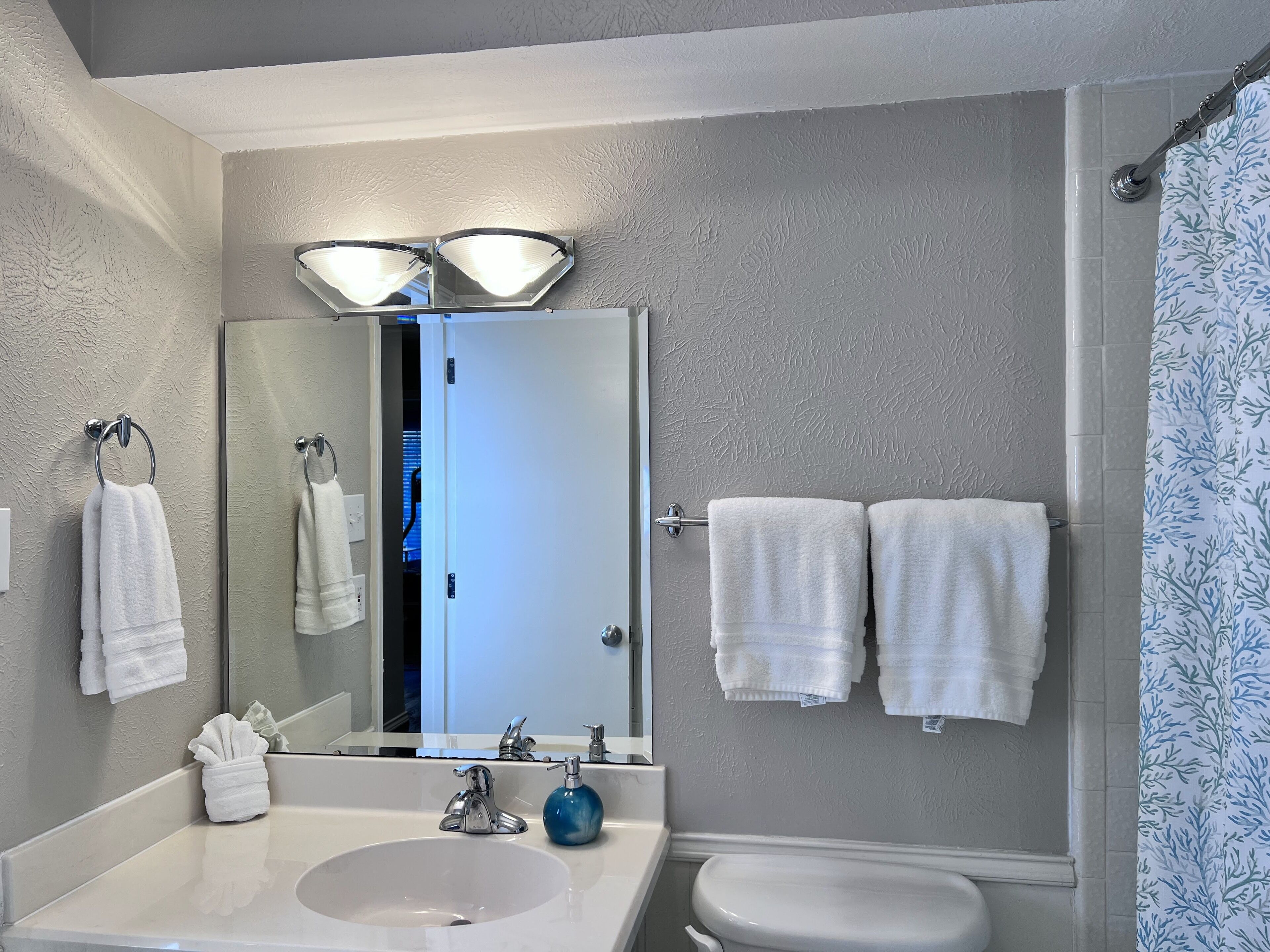 A bathroom with a white sink, a mirror, two overhead lights, a toilet, and a shower curtain with a leaf pattern.
