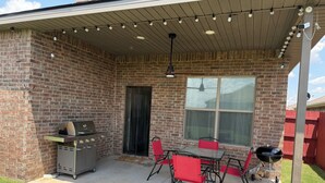 Une terrasse couverte avec un mur en briques, un barbecue, une table avec des chaises et des guirlandes lumineuses.